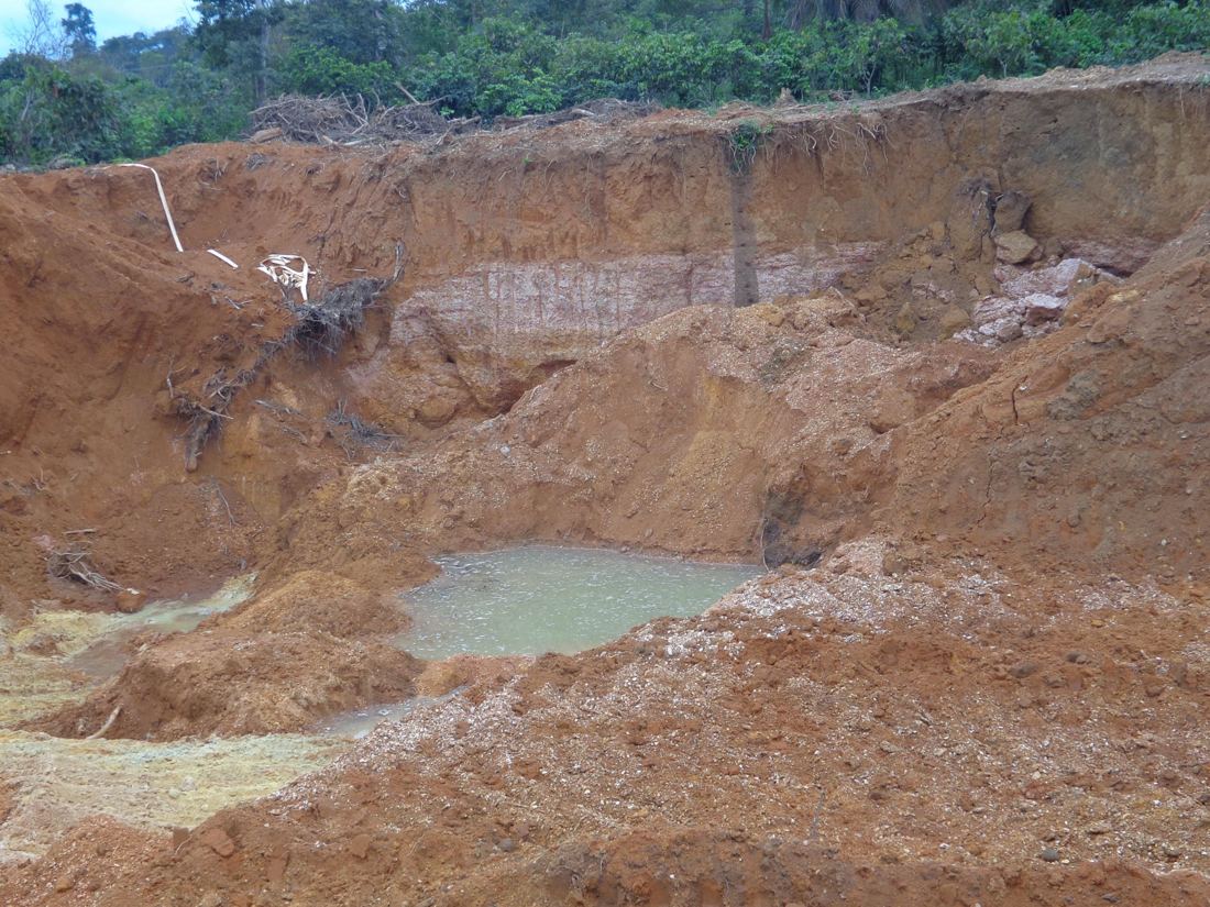 A deep open pit with exposed red soil, uprooted vegetation and a pond of green water