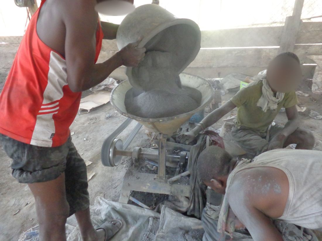Workers feeding material into a grinding machine in a dusty processing shed